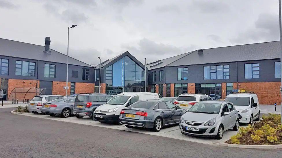 A school building of brick and dark grey walls with cars parked outside and a grey sky overhead