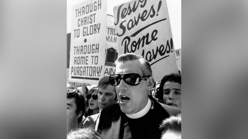 A black and white image of a crowd of protesters. In the centre is Ian Paisley. He is wearing large black sunglasses and a ministers collar. Behind him is a crowd, some are holding placards. One placard says 'Through Christ to Glory - Through Rome to Purgatory'. The other says 'Jesus saves Rome enslaves'. 