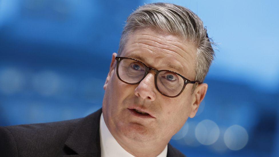 Sir Keir Starmer, with grey hair and glasses, in front of a blue background.  He is visible from the shoulders up and is wearing a black suit and white shirt. 