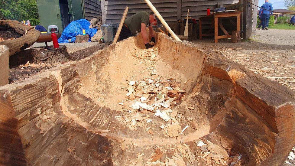 A view of a log from its far end up its chiselled middle towards a man working on carving it out