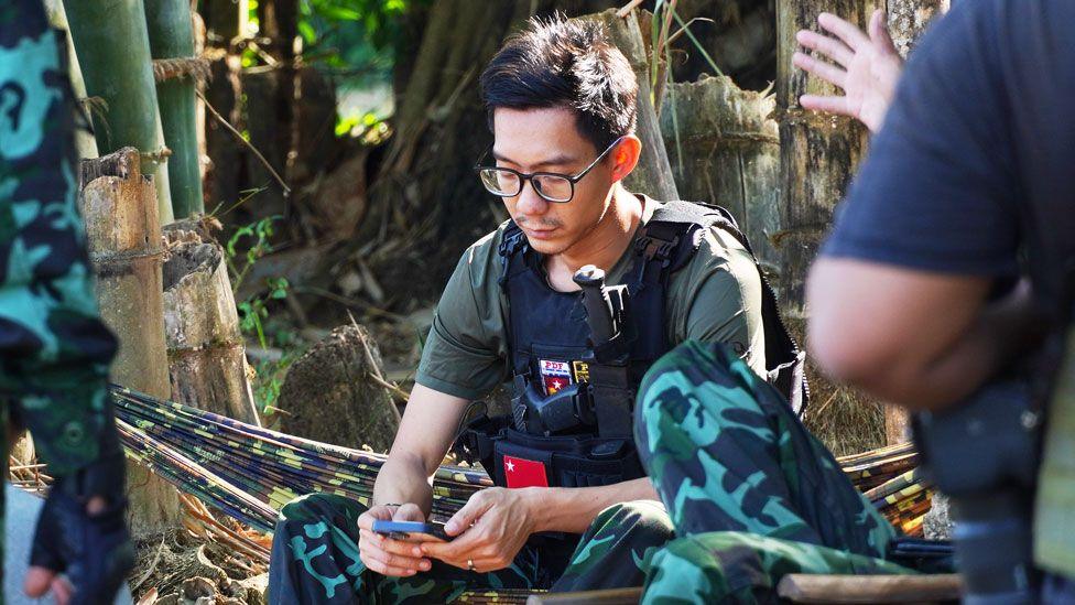 Daeva, wearing a khaki t-shirt and military-style vest, is sitting on a camo-patterned hammock in the forest looking at his phone