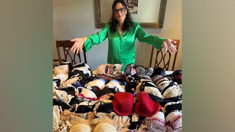 Diana Page showing the bras donated to her. The underwear is on a table in front of her. There is a painting on the wall behind her.