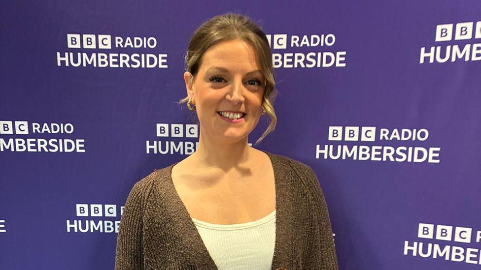 Grace Hunt. She has dark hair and is wearing a white top and brown cardigan. She is standing in front of a purple banner with BBC Radio Humberside logos 