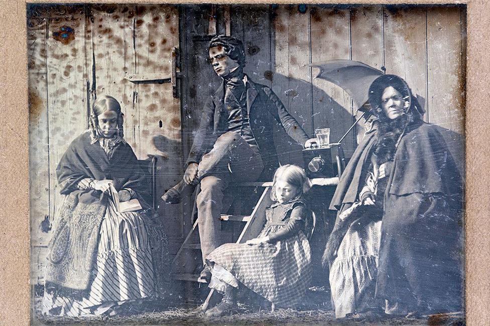 An early black and white image of four figures. Two women and a child are pictured in Victorian clothes, with large skirts, shawls and one is wearing a bonnet. A child wears a similar dress. A gentleman can be seen in a suit.