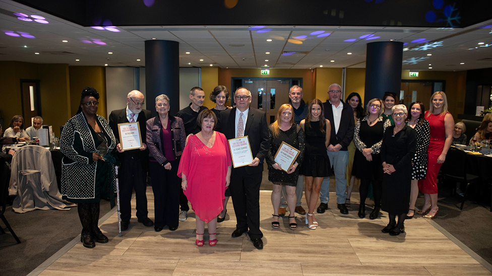 A group of people at an awards evening in a function room, dressed smartly and holding certificates.