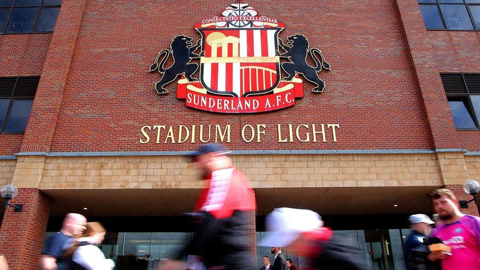 Front entrance to the Stadium of Light in Sunderland with the club's logo on the wall above and the words Stadium of Light written in gold lettering below that. Fans - deliberately blurred by the camera to show motion - are in the foreground.