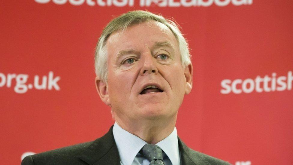 Graeme Pearson photographed against a red backdrop with the Scottish Labour website details in white writing. His mouth is open as if he is speaking. Pearson, who has grey hair combed in a side shed, is wearing a grey suit, light grey shirt and dark grey tie.