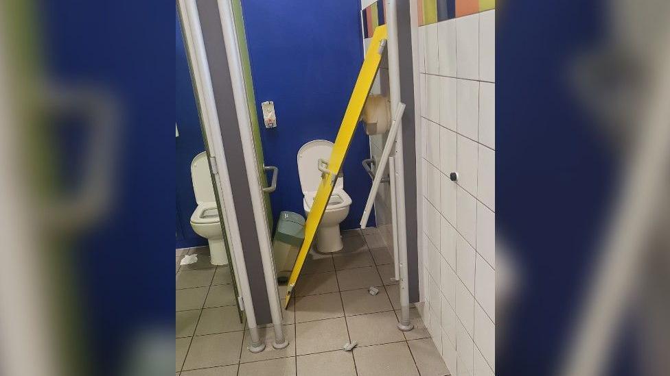 The image shows a yellow toilet door off its hinges and lying diagonal in the toilet cubicle.