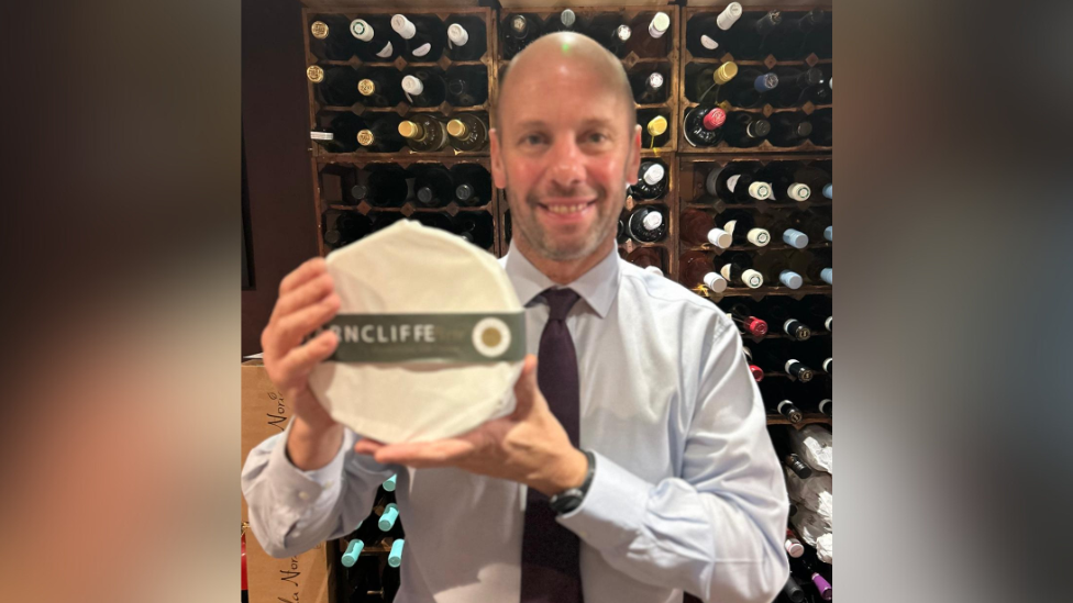 David Straker, dressed in a shirt and tie, holding up a wheel of brie in a wine cellar