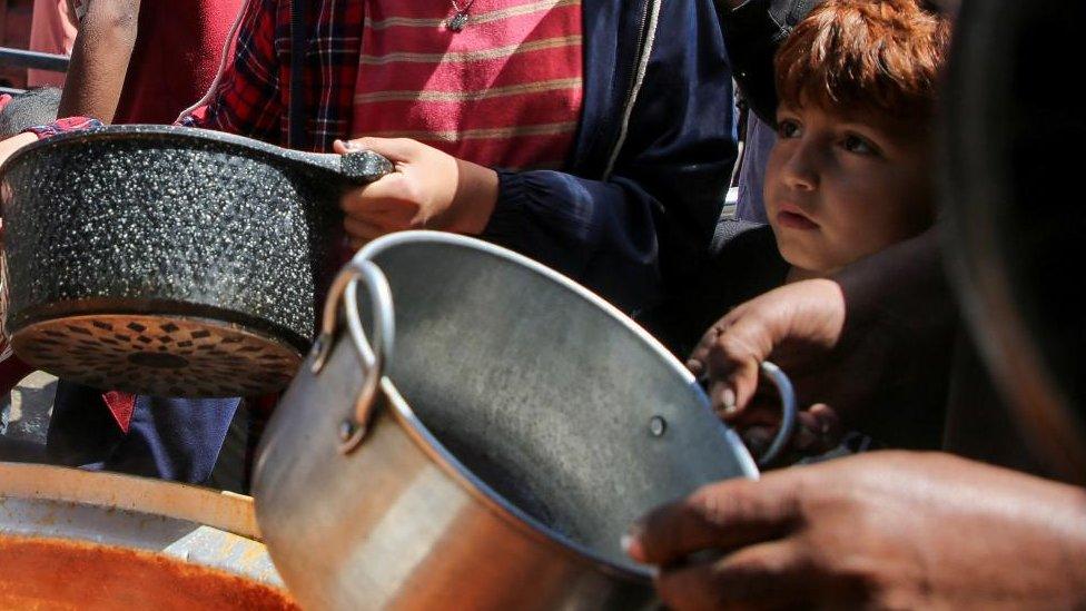 A young boy stands among a crowd holding bowls at an aid kitchen