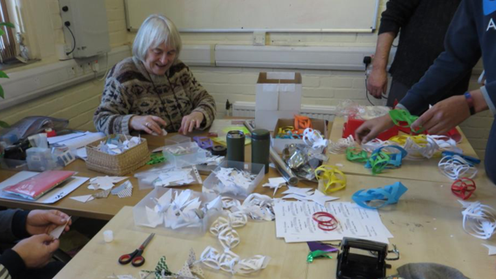 Helen sits at a table filled with coloured paper making origami, with other pairs of hands around her doing the same thing