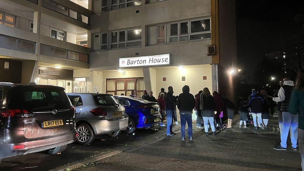 People standing in the dark outside a tower block with lights on