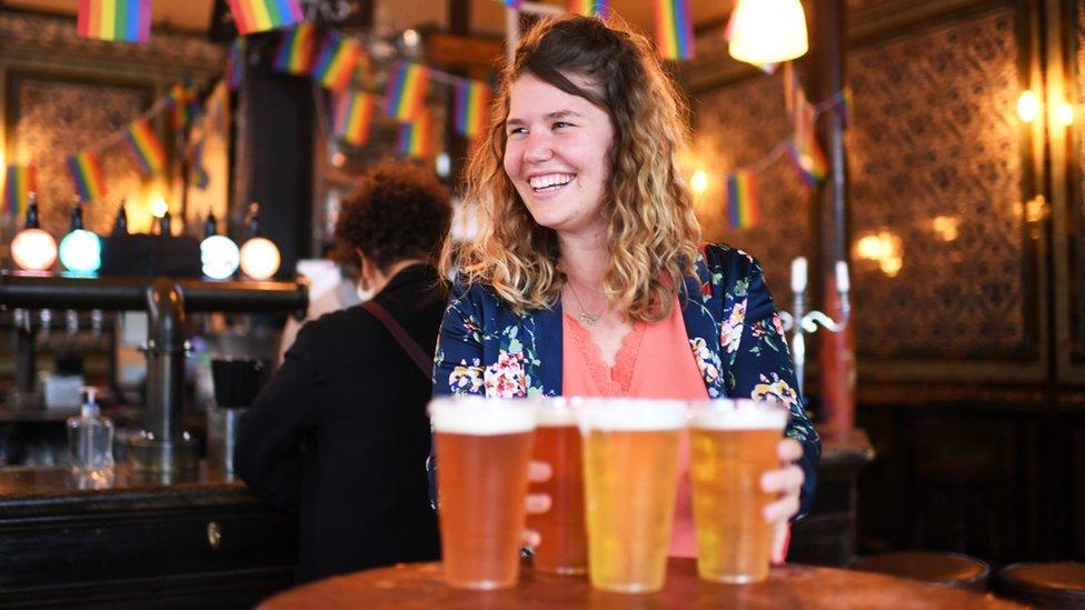 A young woman smiles while holding pints of beer in a pub