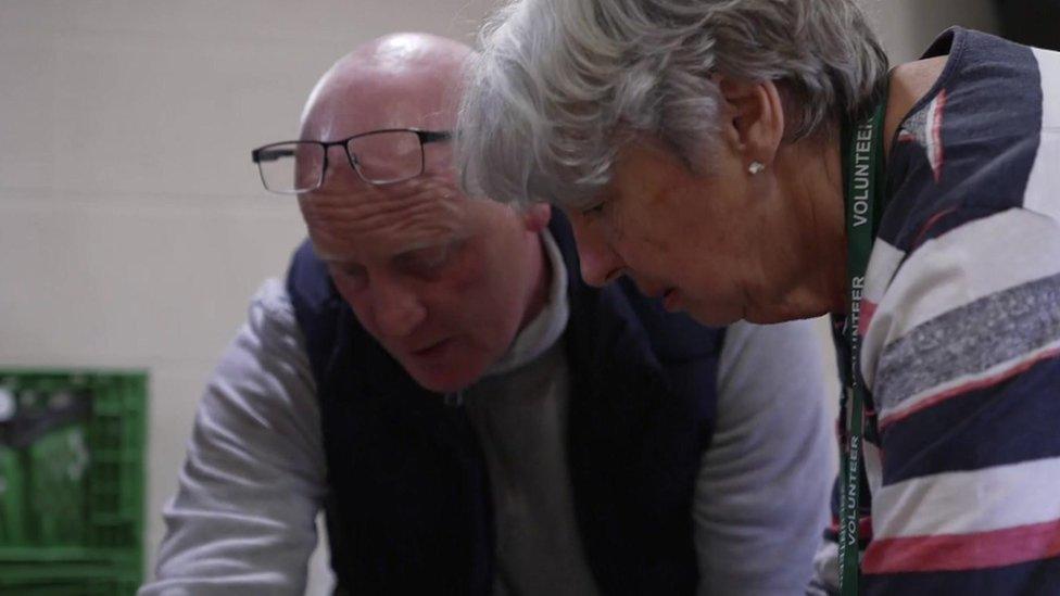 Mr Woodcock talks to a foodbank volunteer while leaning over a crate of food.