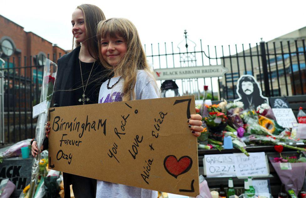 Two girls holding a sign reading "Birmingham Forever Ozzy - You Rock!" in front of the memorial bench with floral tributes