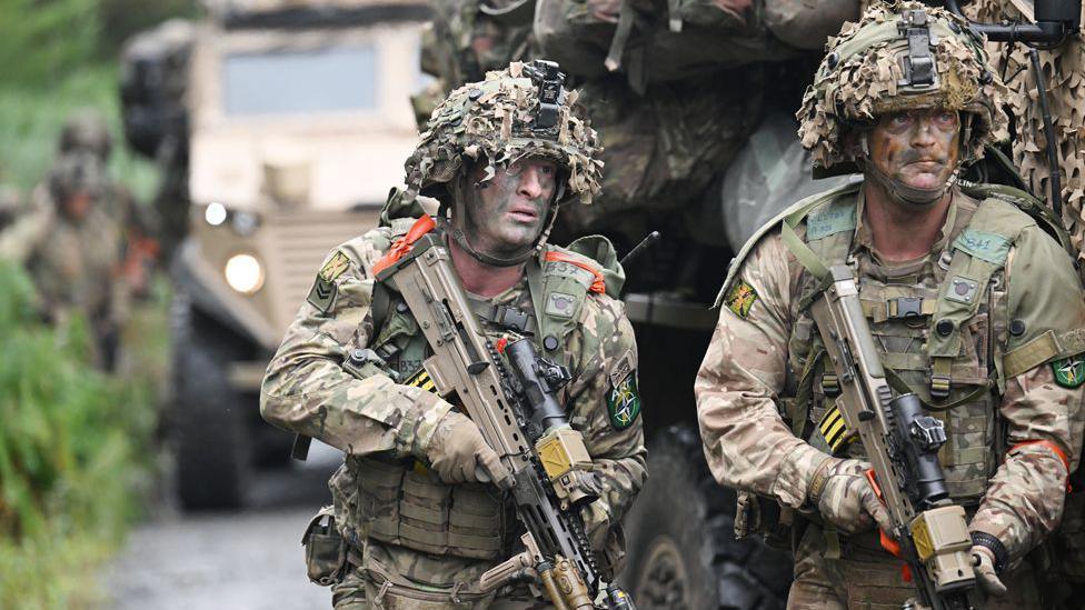 The same two soldiers are now standing side by side carrying rifles and looking towards the right of the photo.