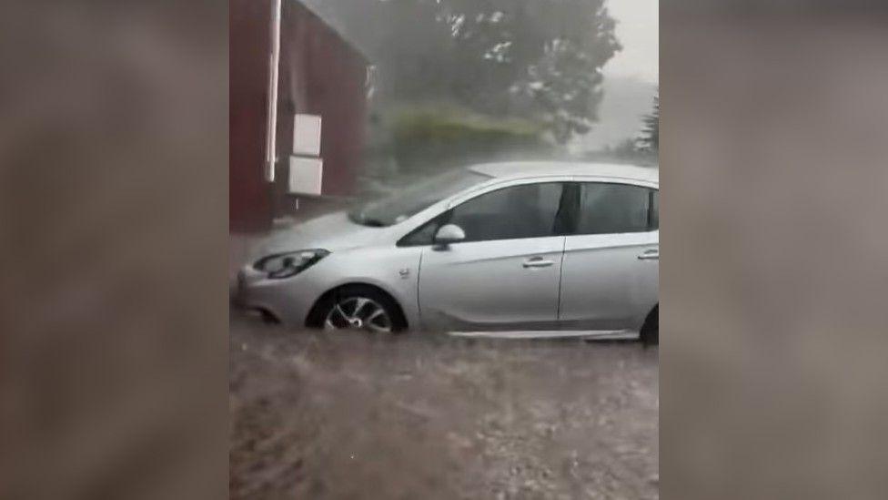 A silver car sits on a flooded driveway as heavy rain falls. A red brick neighbouring property can be seen behind it, as can a hedge and a tree.