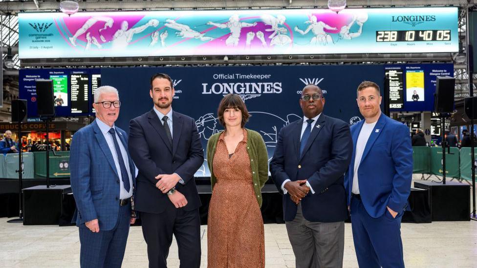 A group of five people, standing in front of a large countdown clock. The group are four men and one woman, Agnes Jones, standing in the centre. She is wearing a tan dress and green cardigan. The four men are all dressed in blue suits. 