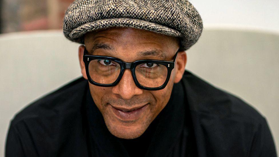 Jay Blades wearing a grey cap, glasses and a dark top, and looking at the camera.