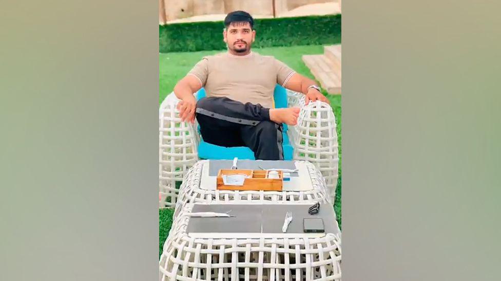 An image showing Fadi Gujjar at a beachside cafe. He is wearing a dark tracksuit and a t-shirt and is sitting on a white chair. In the foreground the table is set for dinner.
