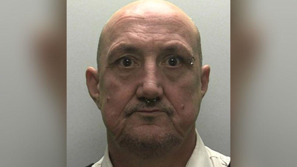 A police mugshot of Simon Mumford. He is a bald man with a septum piercing, an eyebrow piercing on the right side, and light facial hair in the form of a goatee beard. He is wearing a neutral expression.