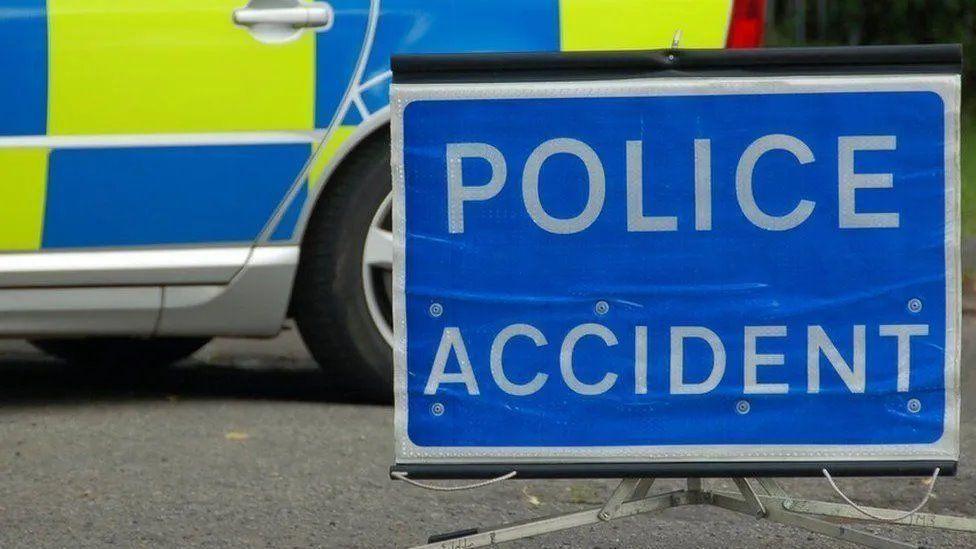 A close up of a blue rectangular sign, resting on tarmac, saying police accident in white letters. Behind it is a close-up of the rear view of a police car, marked with blue and yellow rectangles. 