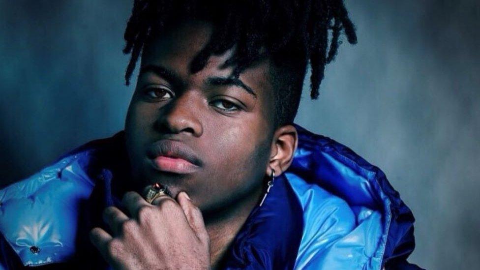 Jordan Adetunji, a black man in his 20s, poses with his hand on his chin and looks at the camera. Jordan has an earring dangling from his left ear and wears a large ring on his left hand as he touches his chin, his braided hair tied up on top of his head. He wears a dark blue puffer jacket with a lighter blue lining showing through. He has dark brown eyes and has a serious expression.