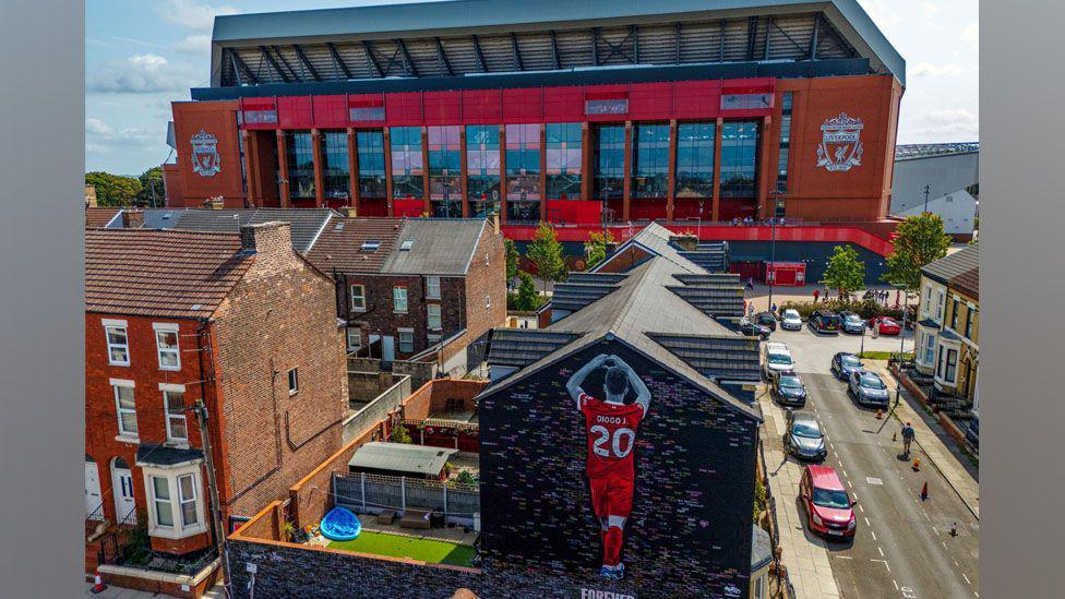 Mural of Jota from the back making a heart shape with his hands. He is wearing a number 20 shirt and red LFC kit against a black background which has has written messages on. Anfield stadium is in the background.