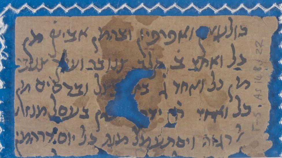 A close-up of a fragment of a medieval manuscript. It has dark writing on a light brown background and is laid on a bright blue background which can be seen through a hole in the manuscript in its middle 