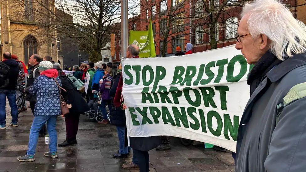 People gathered outside with a man with white hair near the camera in front of a large banner saying 'Stop Bristol Airport Expansion' in green paint.