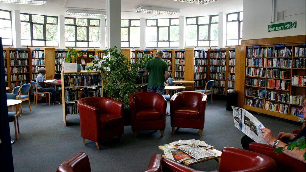 The inside of a library. Shelves of books sit at the edge of the room while tables, red chairs and sofas are placed in the middle. A person sits on a chair reading a newspaper while a standing man in a green top has his back to the camera.