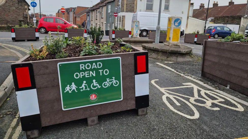 A planter blocks the entrance to a street. It has a sign on it which says 'ROAD OPEN TO' and then has various pictures of people walking, scooting, cycling and travelling in a wheelchair. Next to the planter is a marking of a bike on the road, indicating a cycle lane. 