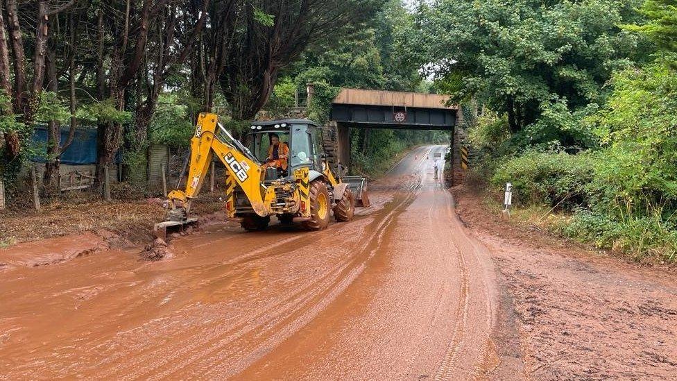 A large yellow digger removing silt and mud from the road. It has cleared a path through the banks of orangey-brown mud which still cover the road.