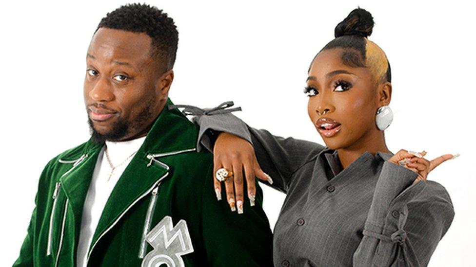 Babatunde and Indiyah on a white background looking at the camera. Babatunde on the left is wearing a white tshirt under a green blazer, holding a silver award which spells out "MOBO". Indiyah on the right has her right arm over his shoulder, and is wearing a grey outfit with silver earrings. Her left hand is doing a "peace" sign.