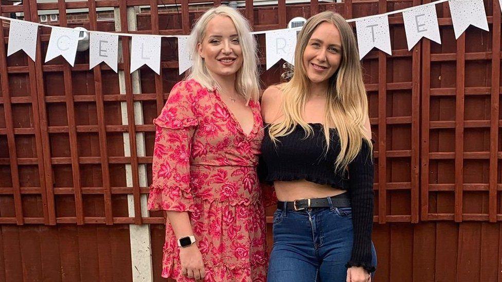 Ashleigh and April stand next to each other with their arms around each other. They are smiling to the camera in front of a fence with a banner hanging on it. Ashleigh has shoulder length white hair and April has long blonde hair.