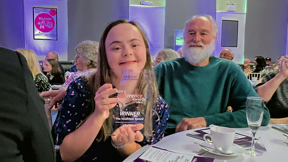 Ellie Taylor sitting at a table next to an older gentleman, holding her award and smiling at the event in Madeley