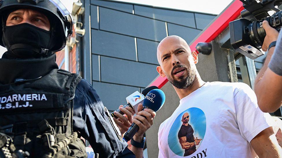 Andrew Tate looks at the camera while being brought in by police, a masked police officer wearing a helmet looks on while reporters gather round Tate, taken outside the Tate brothers' residence in Bucharest on Wednesday night