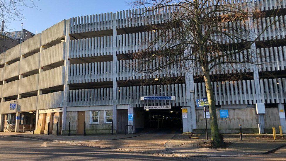 A metal-sided multi-storey car park with entrance at street level and a bare tree on the pavement in front of it