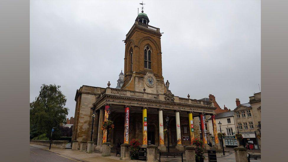 All Saints' Church, Northampton. It is a stone-built late 17th Century building, with pillars at its front, supporting a portico and a tall brick-built tower behind, with a blue-faced clock in its lower level. In front of it is an open space, to its right a row of 19th Century appearing three storey buildings with shops on their ground floor. 