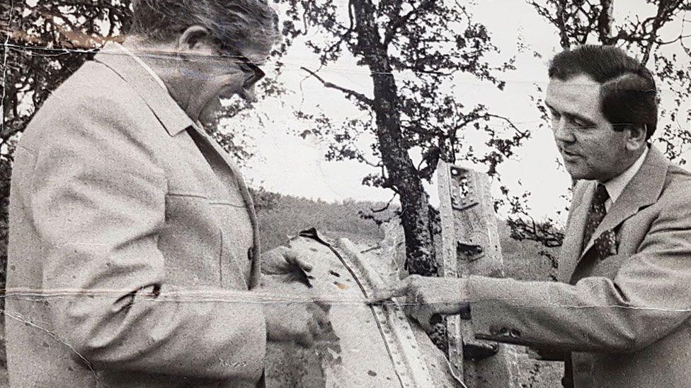 A black and white photo from 1972 showing from the left Einar Thronsen and Michael Hicks holding up a bit of wreckage from the aircraft between them. They have short hair and are wearing jackets. Behind them are trees.
