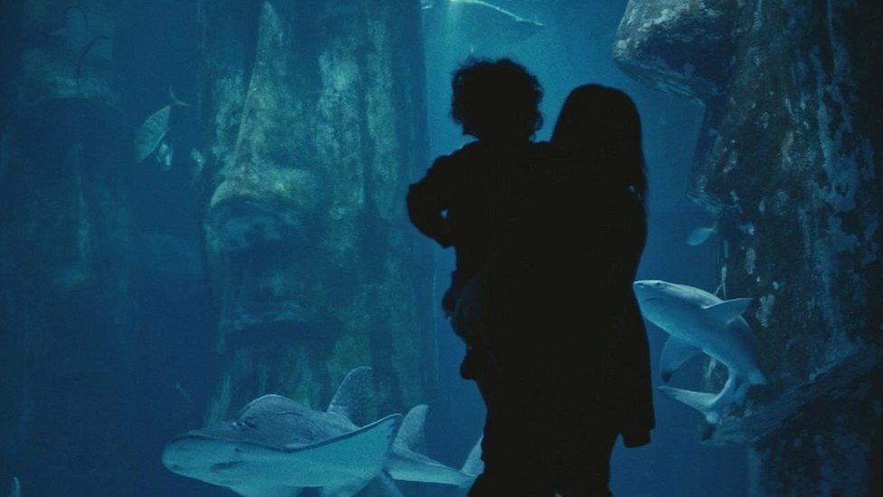 Woman holding child in front of tank of sharks.