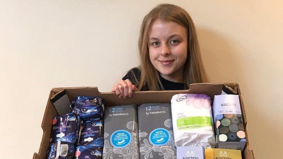 A young woman with long straight hair holds up a carboard box containing a selection of period pads in packages. She's standing against a plain, cream-coloured wall.