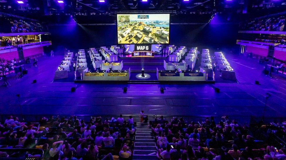 An arena bathed in the purple glow of stage lights. It's set up for a gaming contest - we can see enclosures for each team surrounding a large - Jumbotron-sized - screen showing footage from a round of Call of Duty.