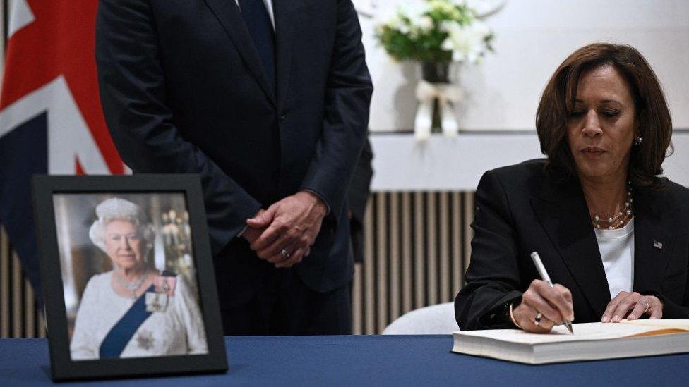 Kamala Harris writing in a large book, image of the queen on her desk