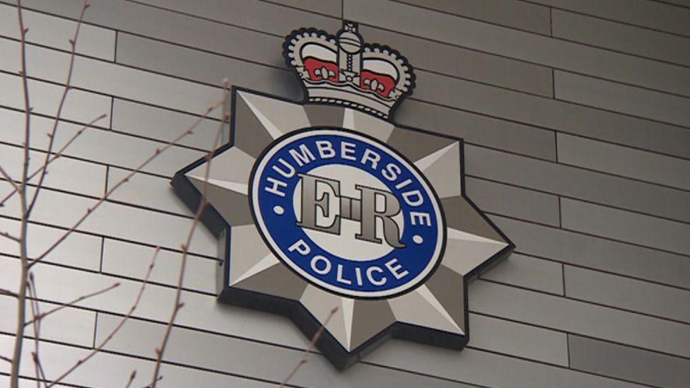 The Humberside Police logo - featuring a blue circle, silver star and red crown - on the side of a building. A tree branch is in the foreground of the image and the building is covered in grey cladding.