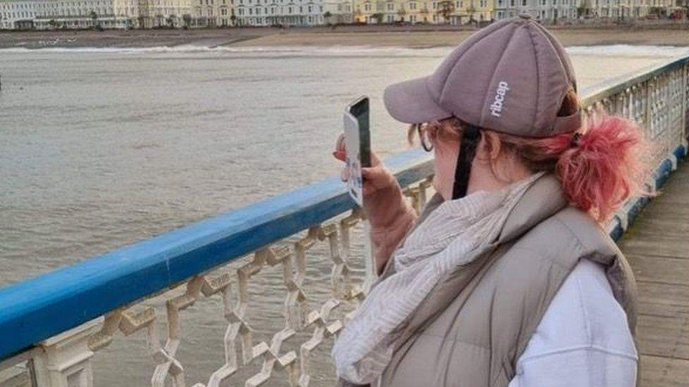 Sophie Price, standing on a pier and holding her phone up. She is wearing a protective cap on her head. In the background you can see the sea, a beach, and a row of houses