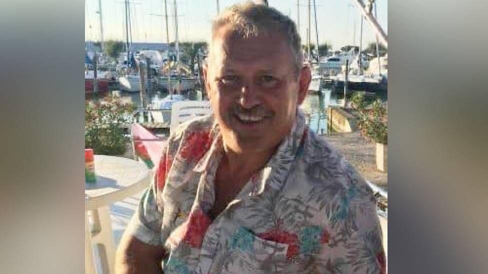 Mike O'Leary wears a tropical shirt, smiling in the sunshine at a harbour, boats visible in the background
