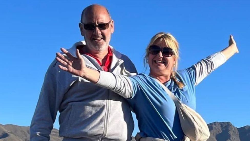 A man and a woman, both smiling and wearing sunglasses and casual clothing, stand smiling in front of some rocky hills. There is a clear blue sky in the background