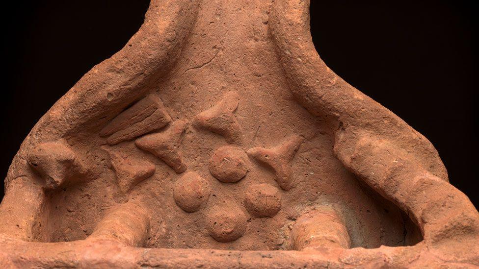 A close-up view of the ground floor of the soul house. It shows low walls on either side, a roughly crafted head of an ox resting on the wall on the left and roughly crafted food items on its floor.