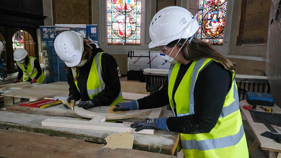 Three apprentices in white hard hats and yellow high vis vests working inside the church on long wooden benches in front of stained glass windows